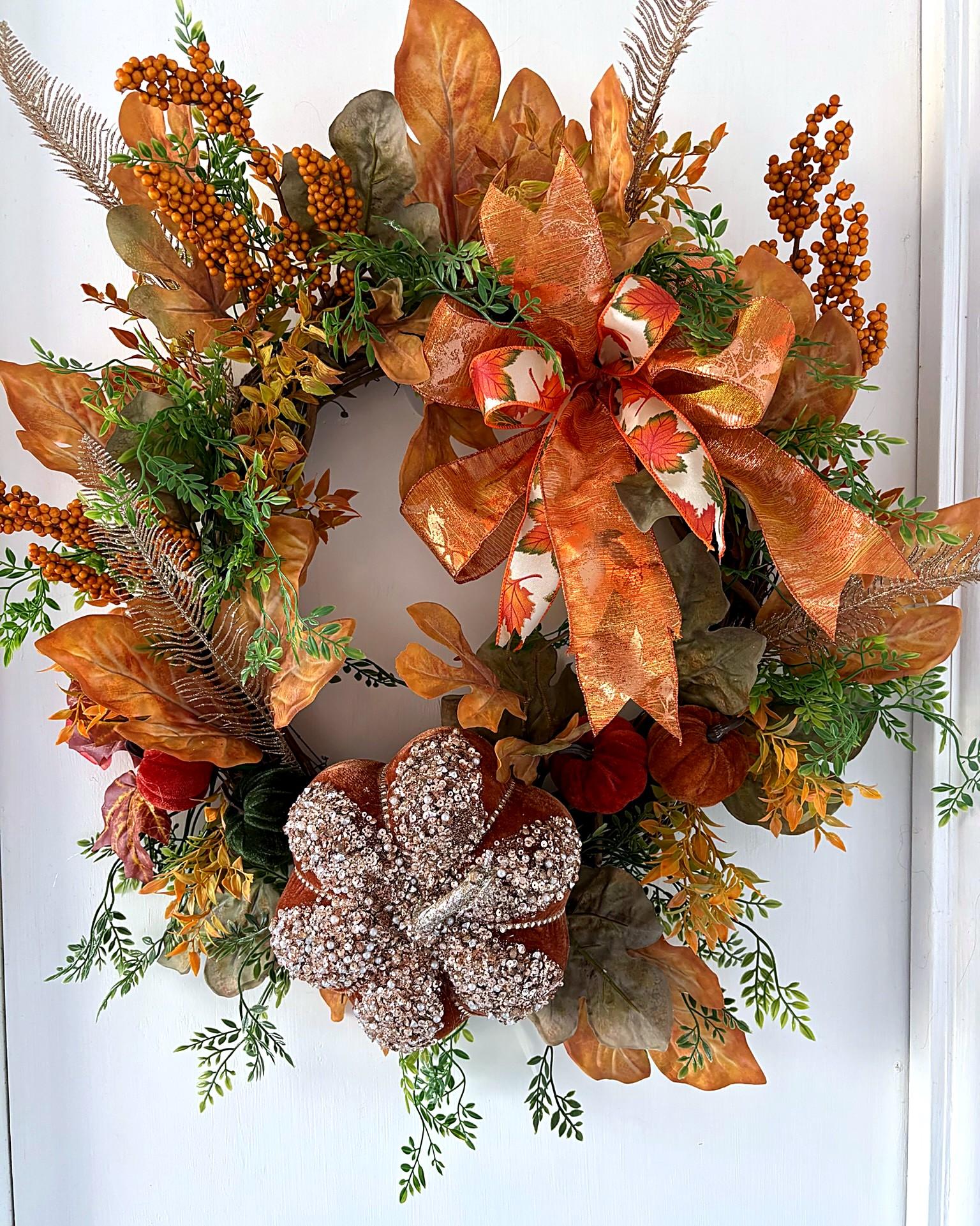 A fall wreath of copper leaves, orange berries and a glittered pumpkin with a copper bow, on a white door