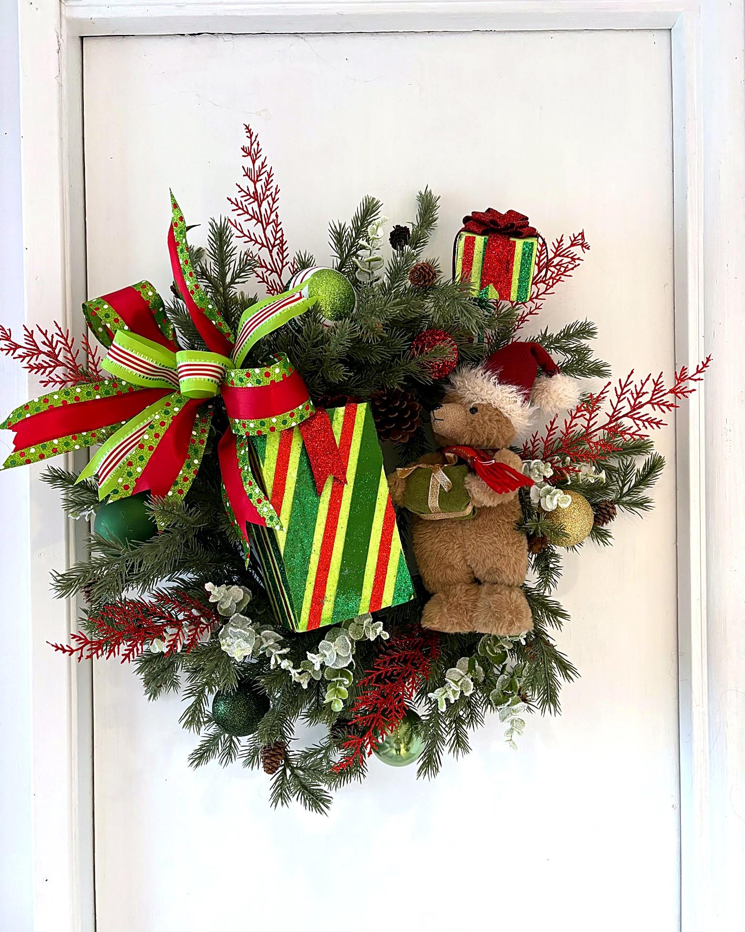 A Christmas greenery wreath with a plush teddy bear in a Santa hat, striped gift boxes and a lime-and-red bow