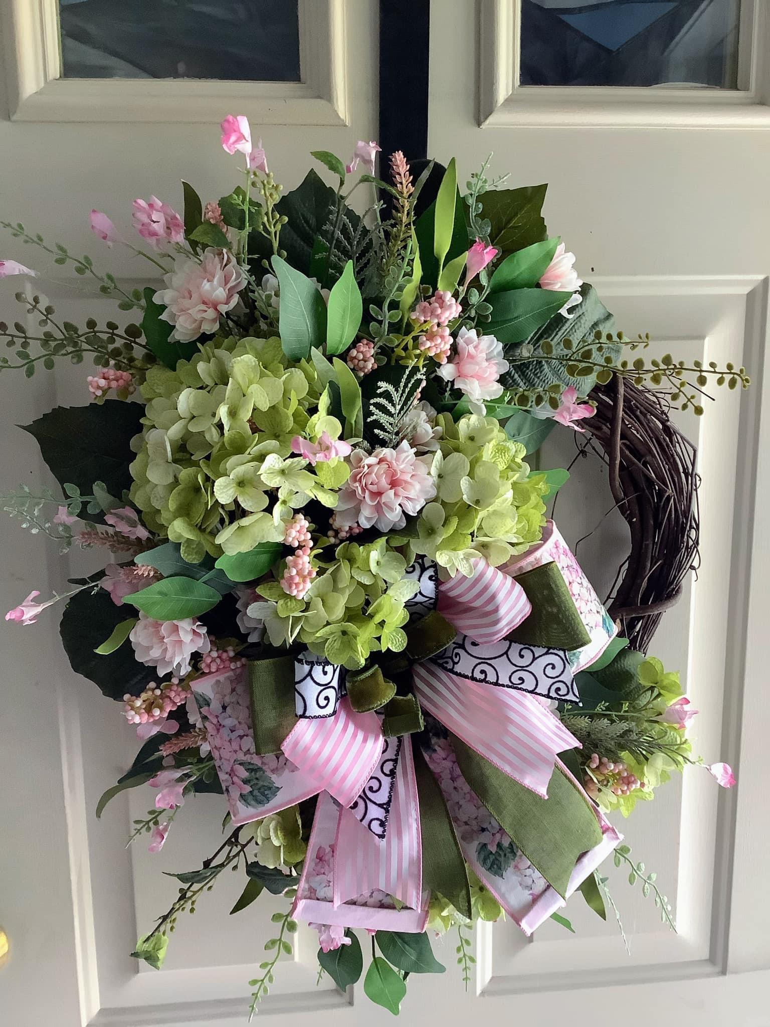 A handmade grapevine wreath with pale green hydrangeas, blush flowers and a pink striped bow, hanging on a white front door